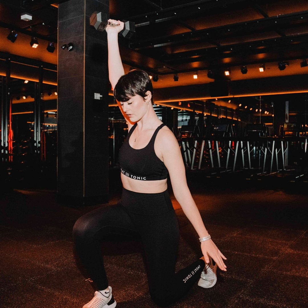 A woman HIIT instructor with short dark hair does a weighted lunge in a neon lit HIIT studio