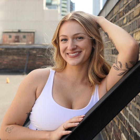 female fitness instructor smiles in a white sports bra white leaning on a beam near a brick wall